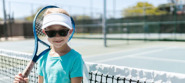 young blonde girl wearing blue holding tennis racket in tennis court wearing black sunglasses