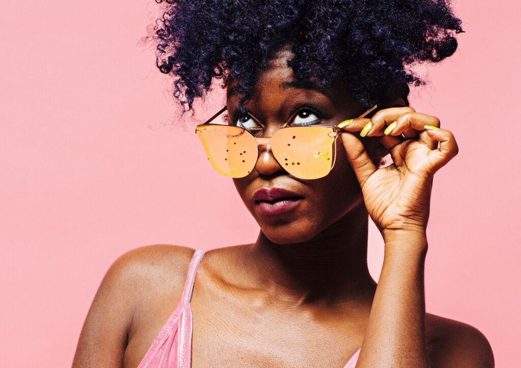 Black woman peeking over mirrored sunglasses with a pink background